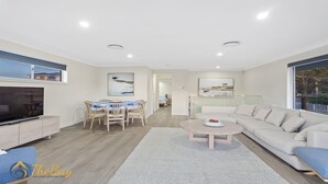 A modern living room with a flat-screen TV, a dining area with a table and chairs, a sofa, and a coffee table. - Twilight on Sunset Beach - Soldiers Point - Amazing sunset views (SOLDIERS POINT)