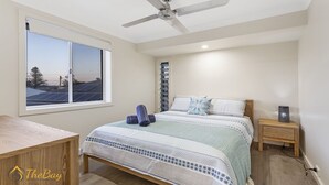 A bedroom with a bed, a desk, a nightstand, a ceiling fan, and a window with a view of solar panels. - Twilight on Sunset Beach - Soldiers Point - Amazing sunset views (SOLDIERS POINT)