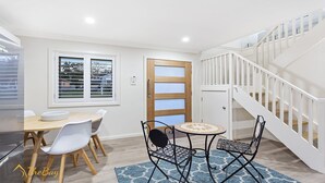 A dining area with a wooden table and chairs, a small round table with a patterned table runner, and a staircase with white railings. - Twilight on Sunset Beach - Soldiers Point - Amazing sunset views (SOLDIERS POINT)
