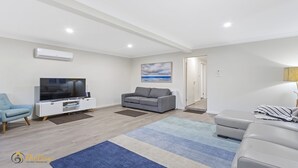 A modern living room with a flat-screen TV, grey sofa, blue armchair, and a large rug. - Twilight on Sunset Beach - Soldiers Point - Amazing sunset views (SOLDIERS POINT)