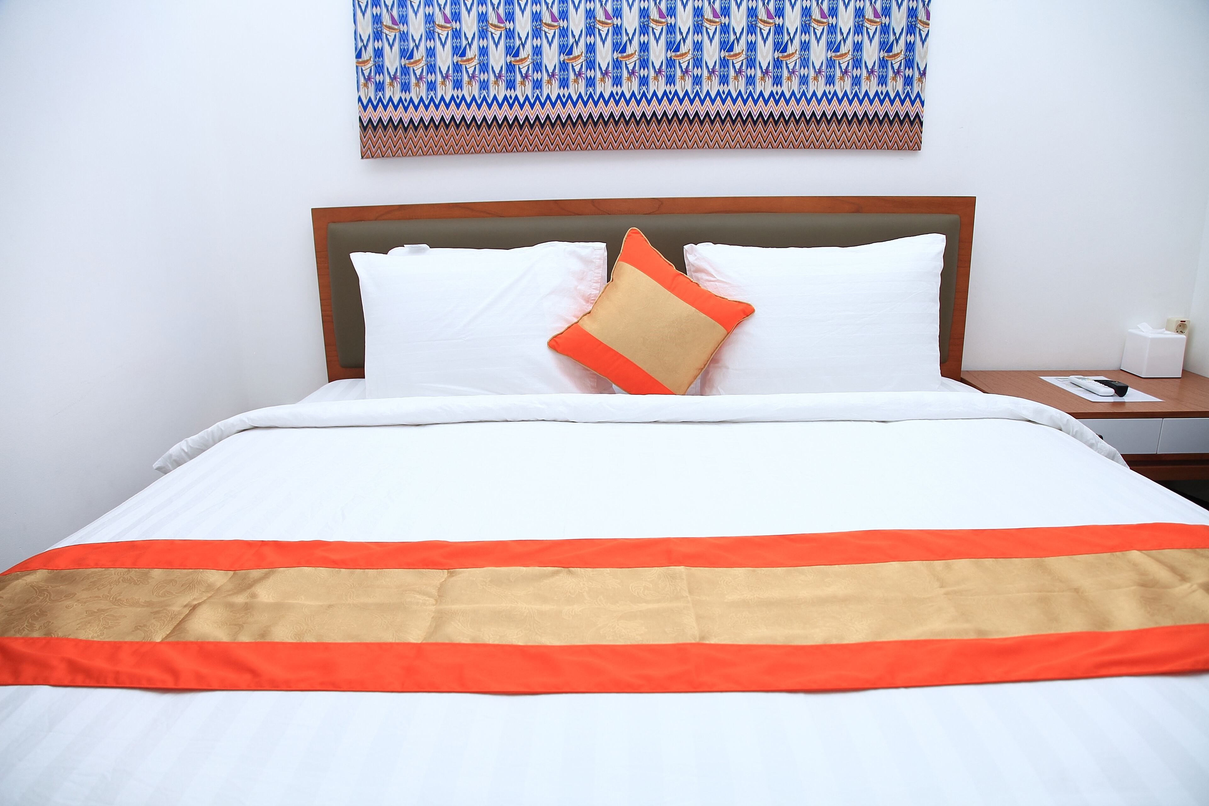 A neatly made bed with white and orange bedding, a patterned wall hanging above, and a bedside table with a tissue box.