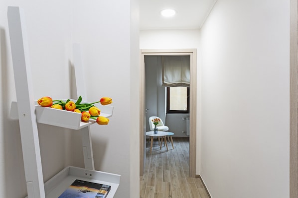 A hallway with a white wall, a white shelf with yellow tulips, a white chair, and a small table with a vase of flowers.
