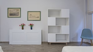 Una habitación con una consola blanca, dos cuadros enmarcados, un estante blanco y una silla azul.