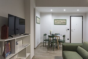 A compact living space with a flat-screen TV on a white shelf, a dining table with chairs, a green sofa, and a door with a framed picture above it.