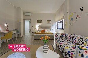 A modern hotel room with a bed, a desk, a chair, a sofa, and a small table with a vase of flowers.