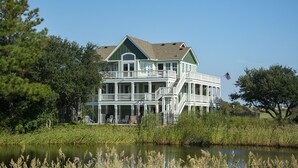 A large, multi-story house with a green roof and white exterior, surrounded by trees and a body of water. - Brown Dog by the Sea: Soundside, Private Pool, Spa (Corolla)