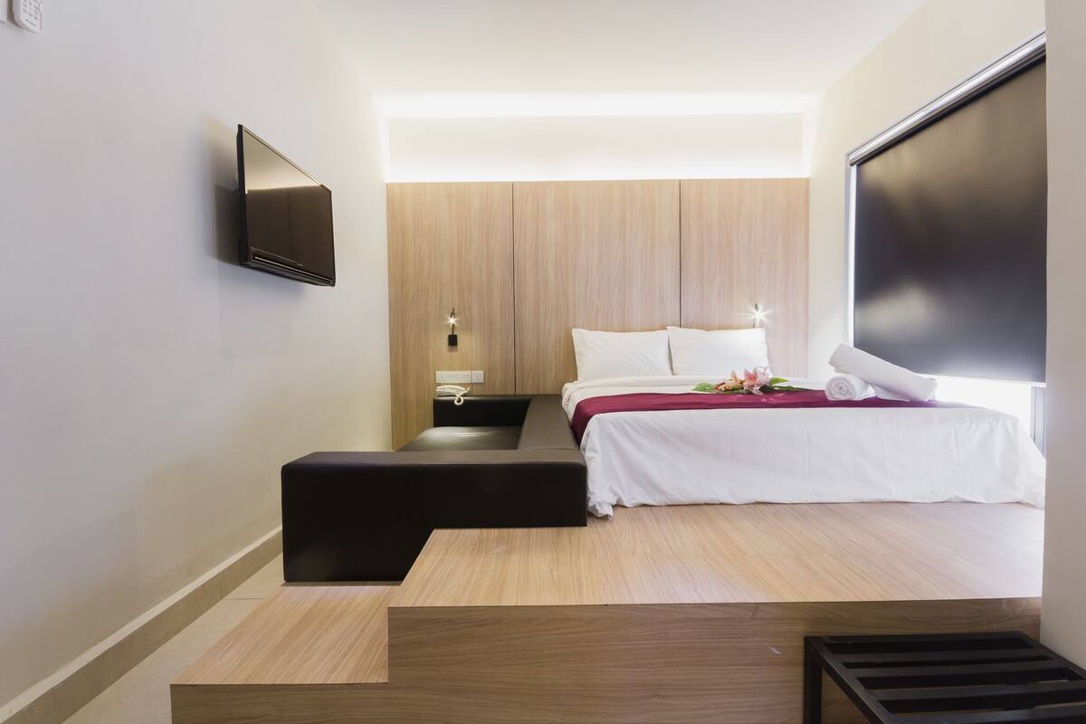 A modern hotel room with a bed, a flat-screen TV mounted on the wall, a bedside table, and a small seating area.