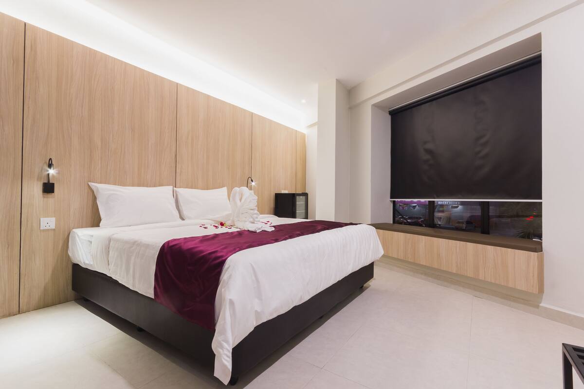A modern bedroom with a large bed, wooden panel walls, a built-in TV, and a window with a view of the city.