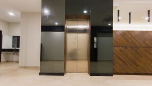 Elevador em edifício moderno com acabamento metálico e painel de madeira à direita.