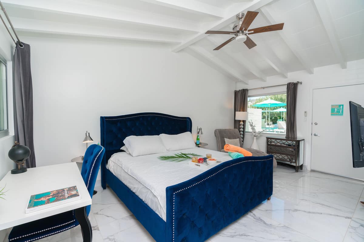 A bedroom with a large bed, a desk, a chair, a ceiling fan, and a view of the outdoors.