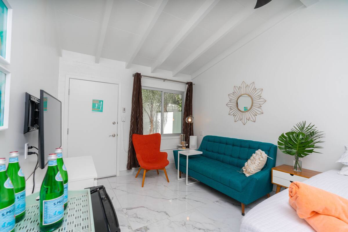 A modern hotel room with a teal sofa, a bright orange chair, a small white table, and a TV mounted on the wall.