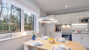 Uma cozinha moderna com armários brancos, uma mesa de jantar de madeira para quatro pessoas e uma grande janela com vista para uma floresta.