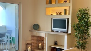 A living room with a TV, a fireplace, a plant, and a dining area visible through a glass door. - Charming Apartment with Private Garden & Pool (Vilamoura)