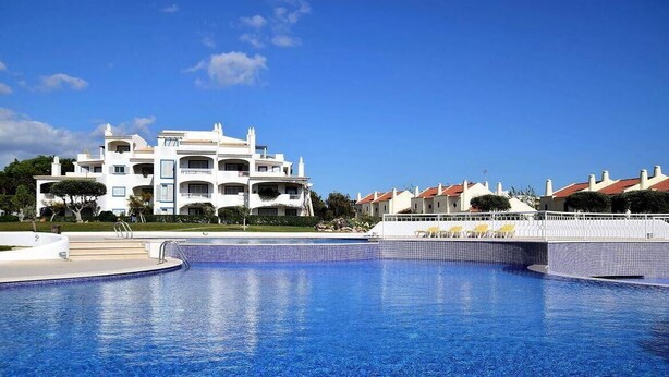 A large swimming pool with a clear blue water surface, surrounded by a white fence and a building with multiple balconies and a red-tiled roof in the background. - Charming Apartment with Private Garden & Pool (Vilamoura)