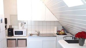 A modern kitchen with white cabinets, a microwave, a coffee maker, and a sink. There are storage boxes under the cabinets and a small table with a chair.