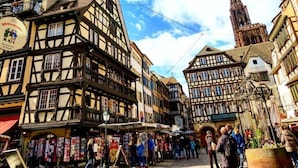 A traditional European town square with half-timbered buildings, a prominent church tower, and a market area with people and stalls.