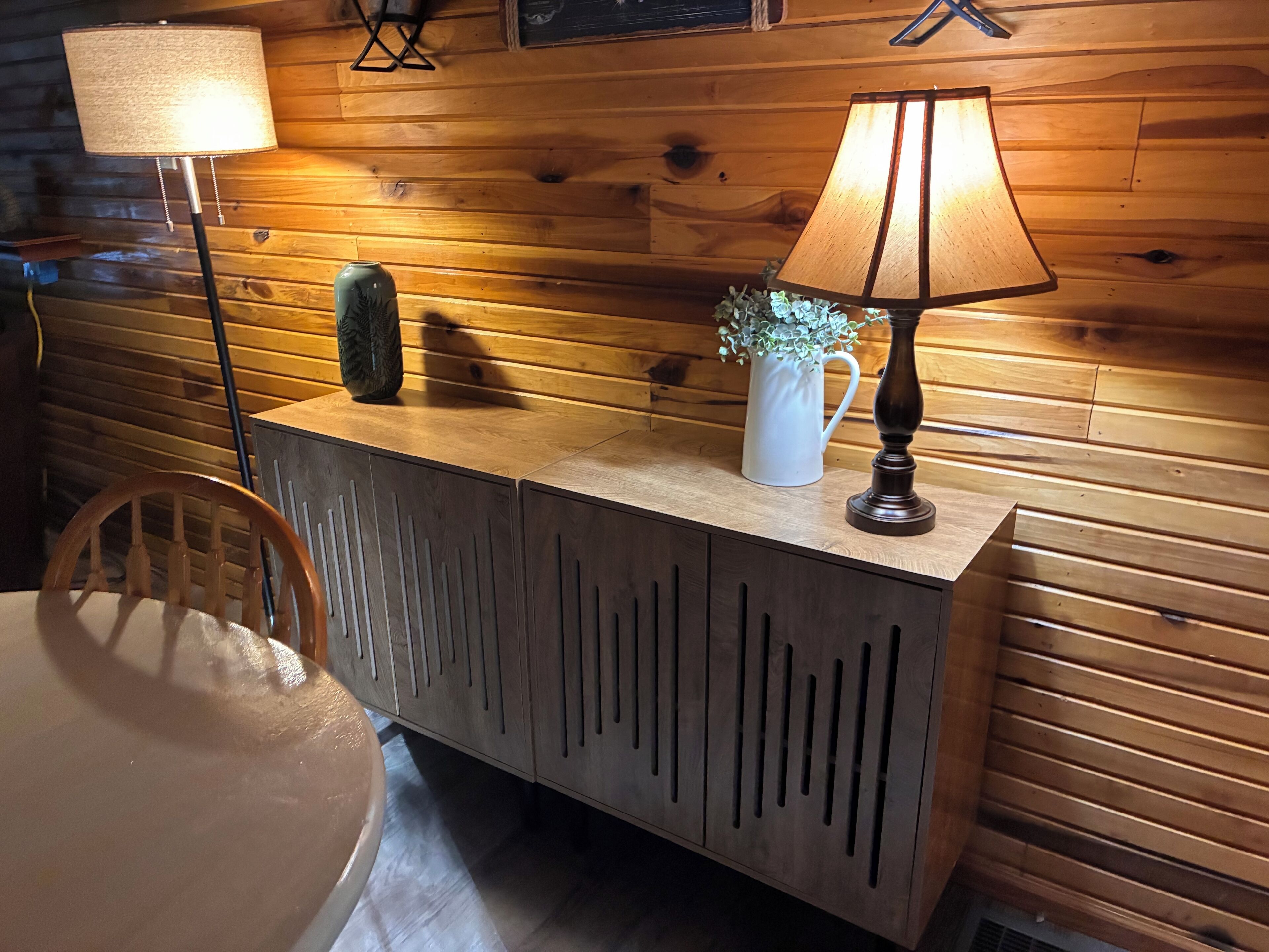 A wooden cabin interior with a wooden cabinet, a lamp, a vase with a plant, and a chair.