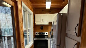 A compact kitchen with wooden walls, a built-in oven, a refrigerator, and a window overlooking a deck.