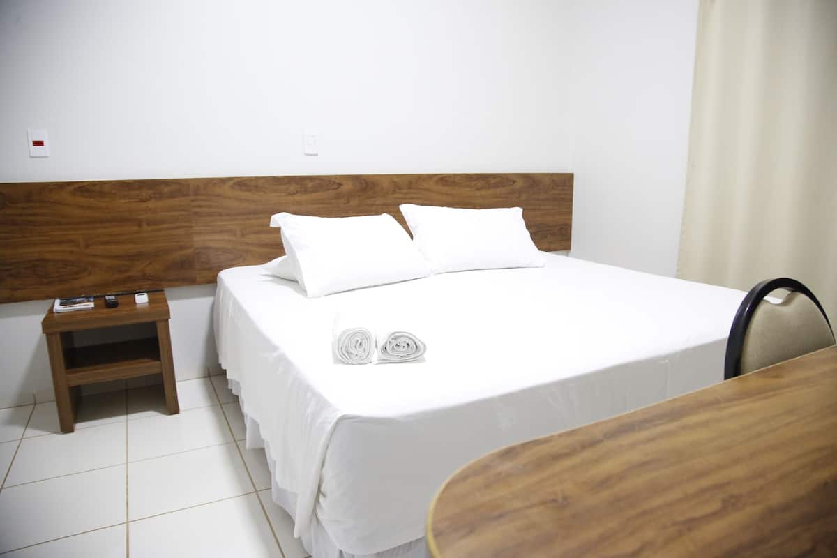 A hotel room with a wooden headboard, a bed with white bedding, a wooden nightstand with a remote control, and a chair.