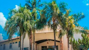 A hotel with a white facade, multiple palm trees in front, and a clear blue sky. - Terras Hotel (Lucas do Rio Verde)