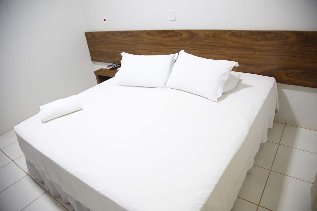 A neatly made bed with white linens and pillows, a wooden headboard, and a white nightstand with a remote control.