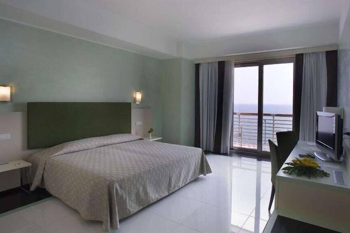 Habitació d'hotel amb un llit, taules de nit, escriptori i un ordinador, i vista al mar.