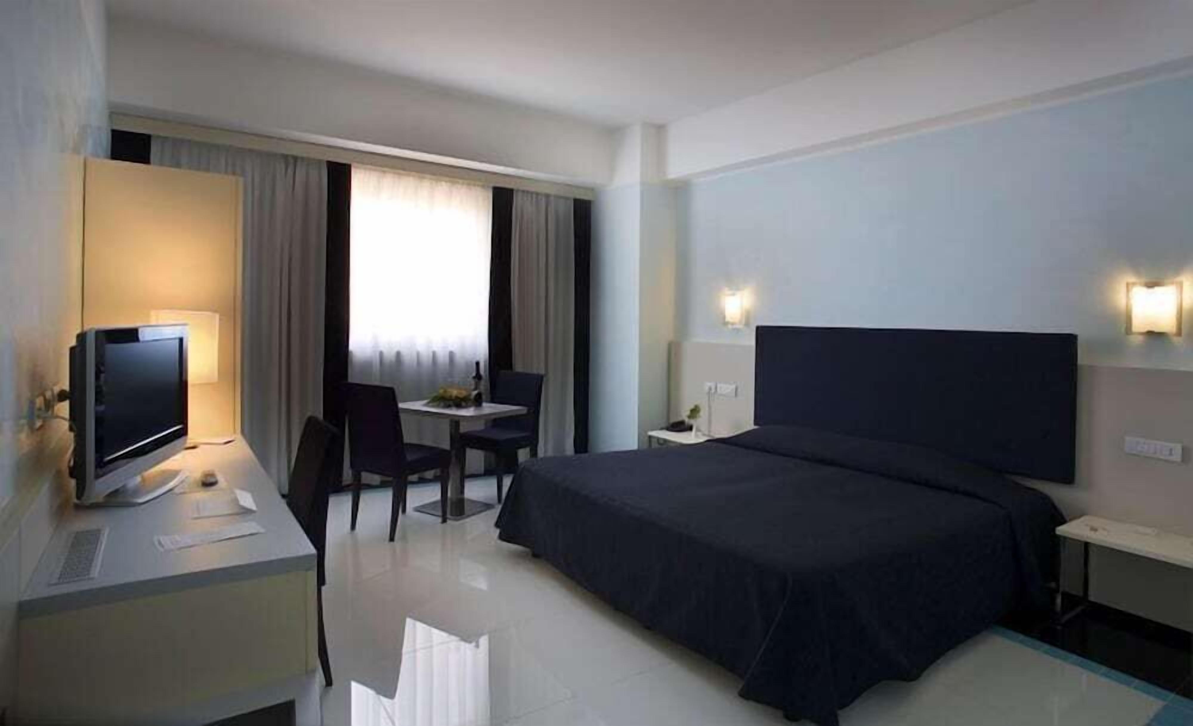 A hotel room with a bed, a desk with a computer, a chair, a small table with a lamp, and a window with curtains.