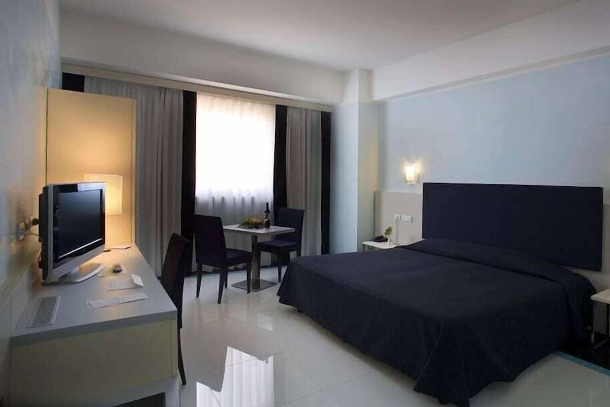 Habitació d'hotel amb un llit, escriptori amb ordinador, cadira, taula petita i finestra amb cortines.