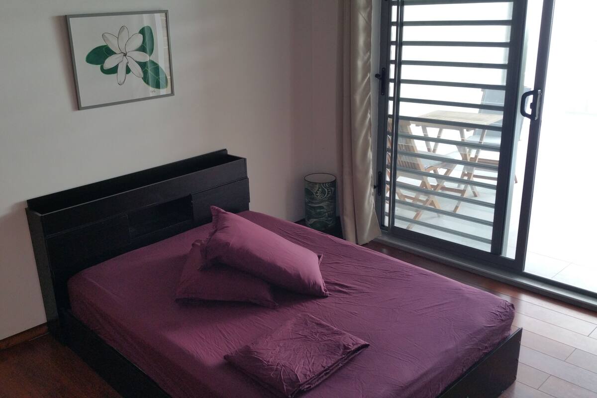 Een slaapkamer met een bed, nachtkastje, een raam met jaloezieën en een aan de muur hangend schilderij.