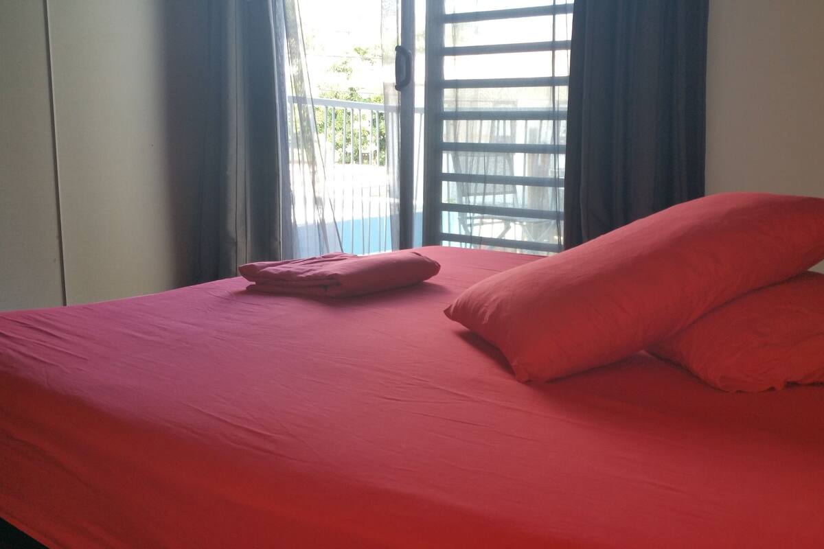 Een bed met rode beddengoed en kussens, een raam met jaloezieën en uitzicht op groen buiten.