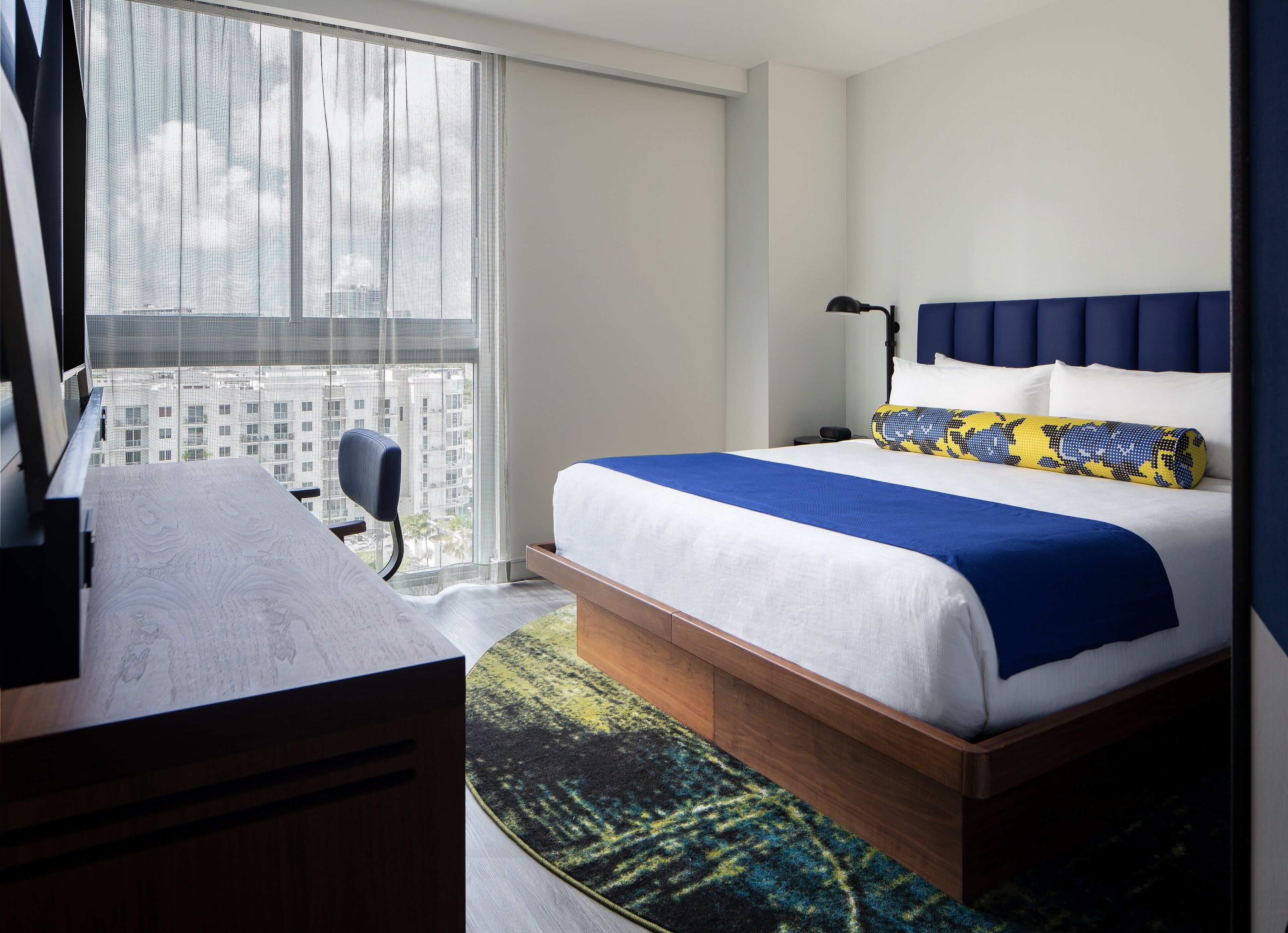 A hotel room with a bed, a desk, a chair, and a large window with a city view.