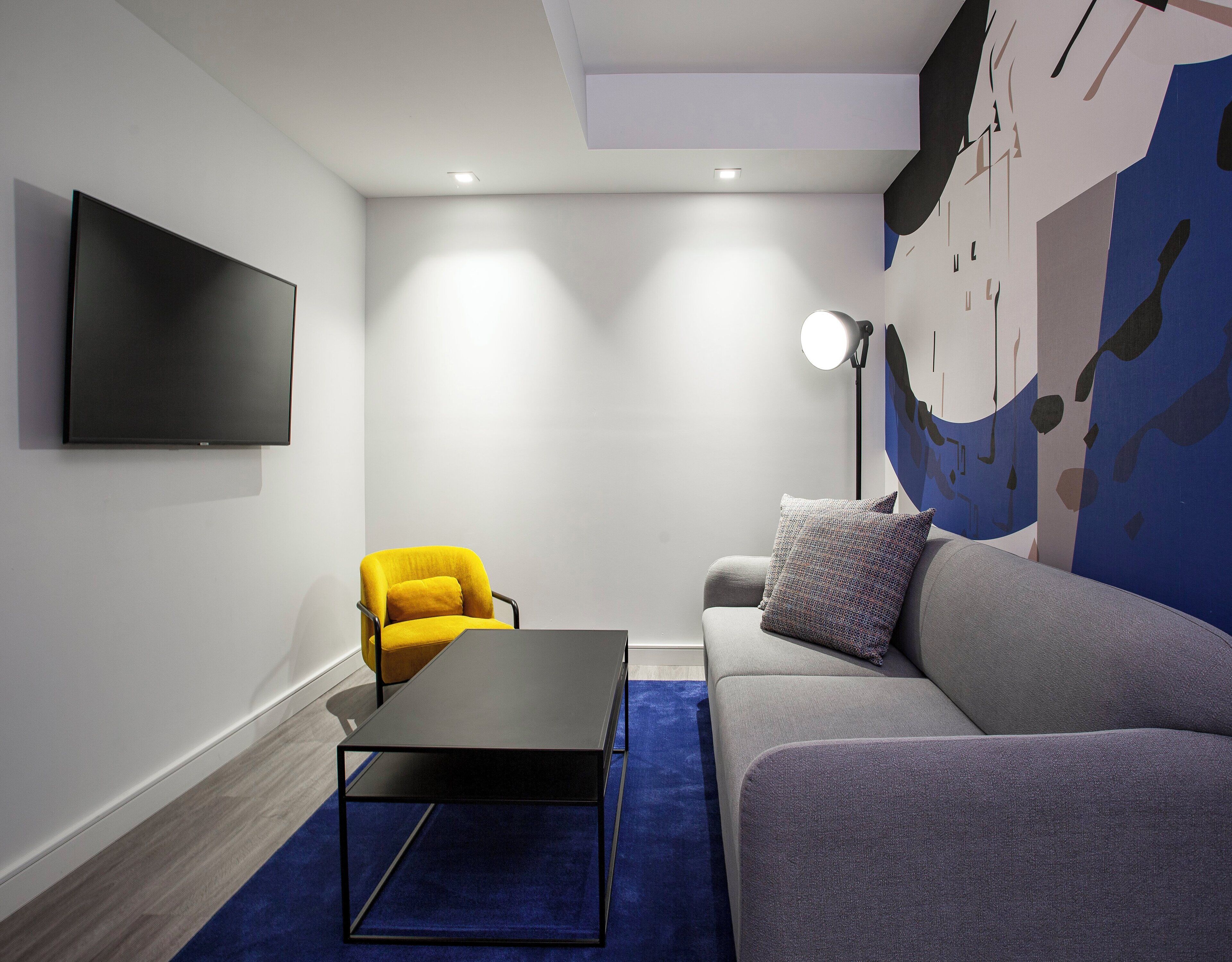 Een moderne woonkamer met een flatscreen televisie, een gele fauteuil, een grijze bank, een zwarte salontafel en een blauw vloerkleed.