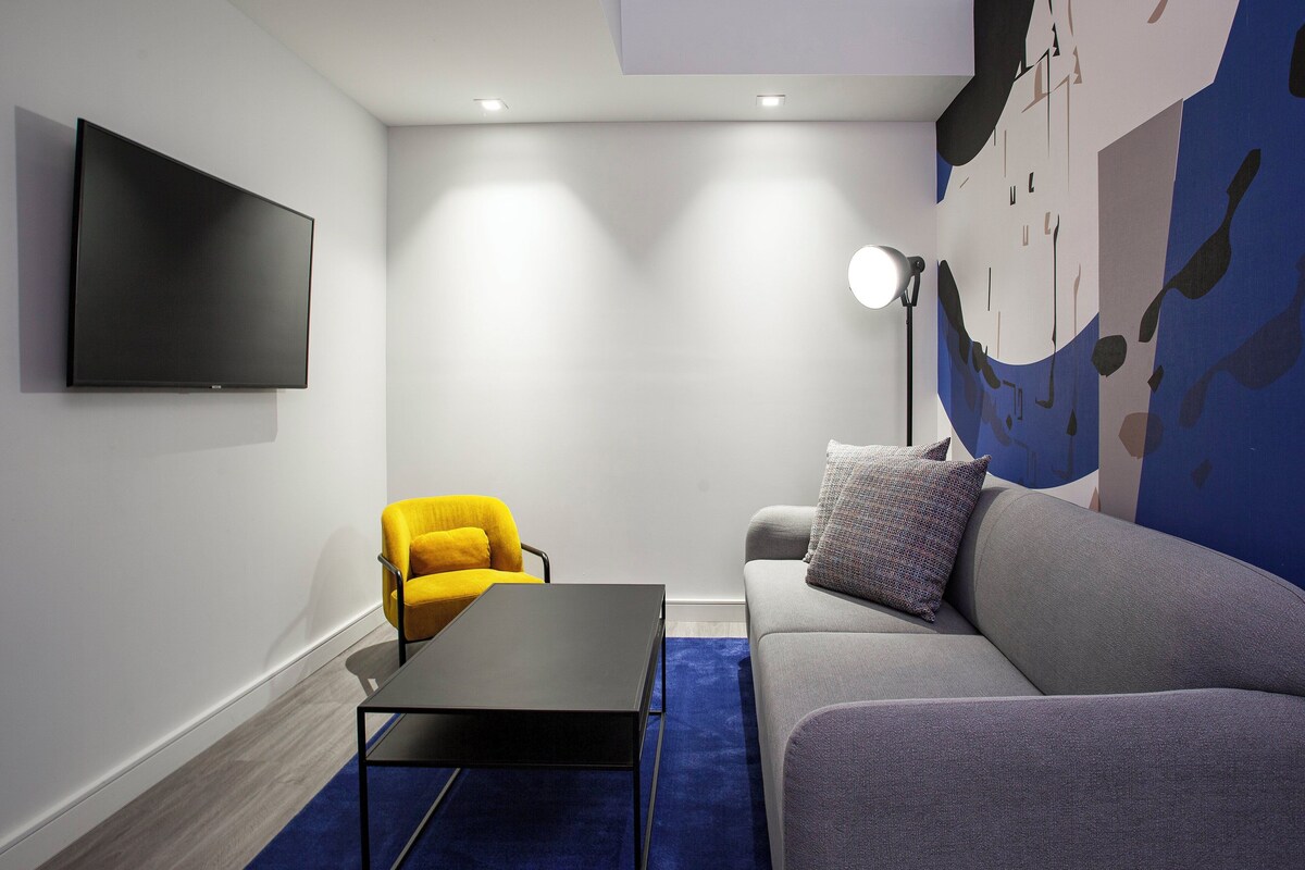 A modern living room with a flat-screen TV, a yellow armchair, a grey sofa, a black coffee table, and a blue rug.