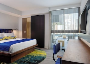 A hotel room with a bed, a desk, a chair, a large window with a view of buildings, and a door leading to a bathroom. - Hotel Indigo Miami Brickell by IHG (Miami)