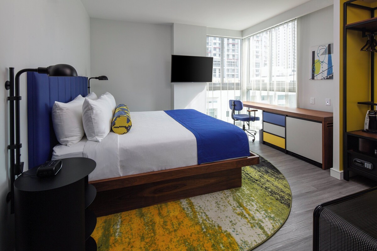 A hotel room with a bed, a desk, a chair, a TV, and a view of the city.