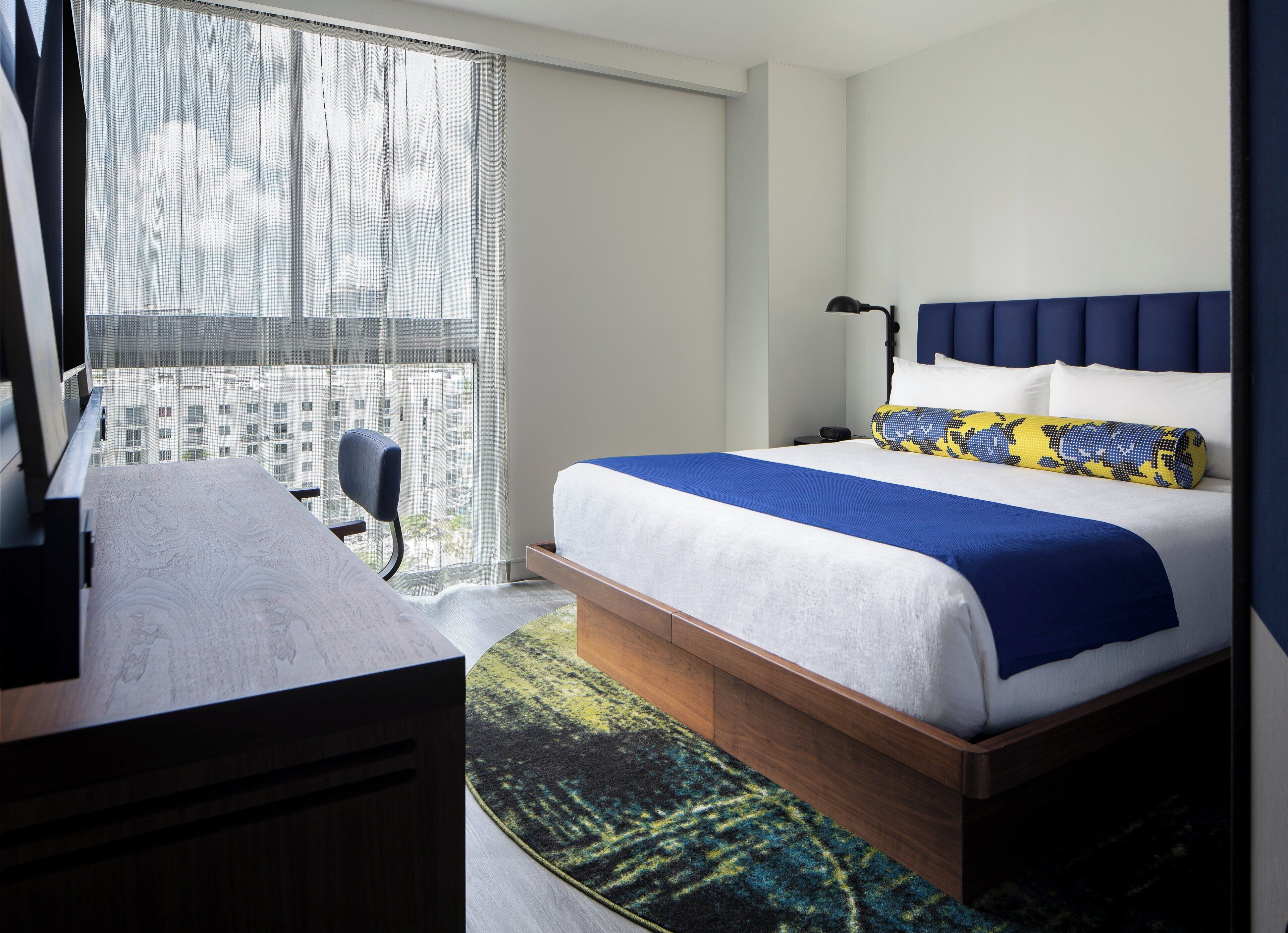 Habitación de hotel con cama, escritorio, silla, ventana con vista a edificios y mesita de noche con lámpara.