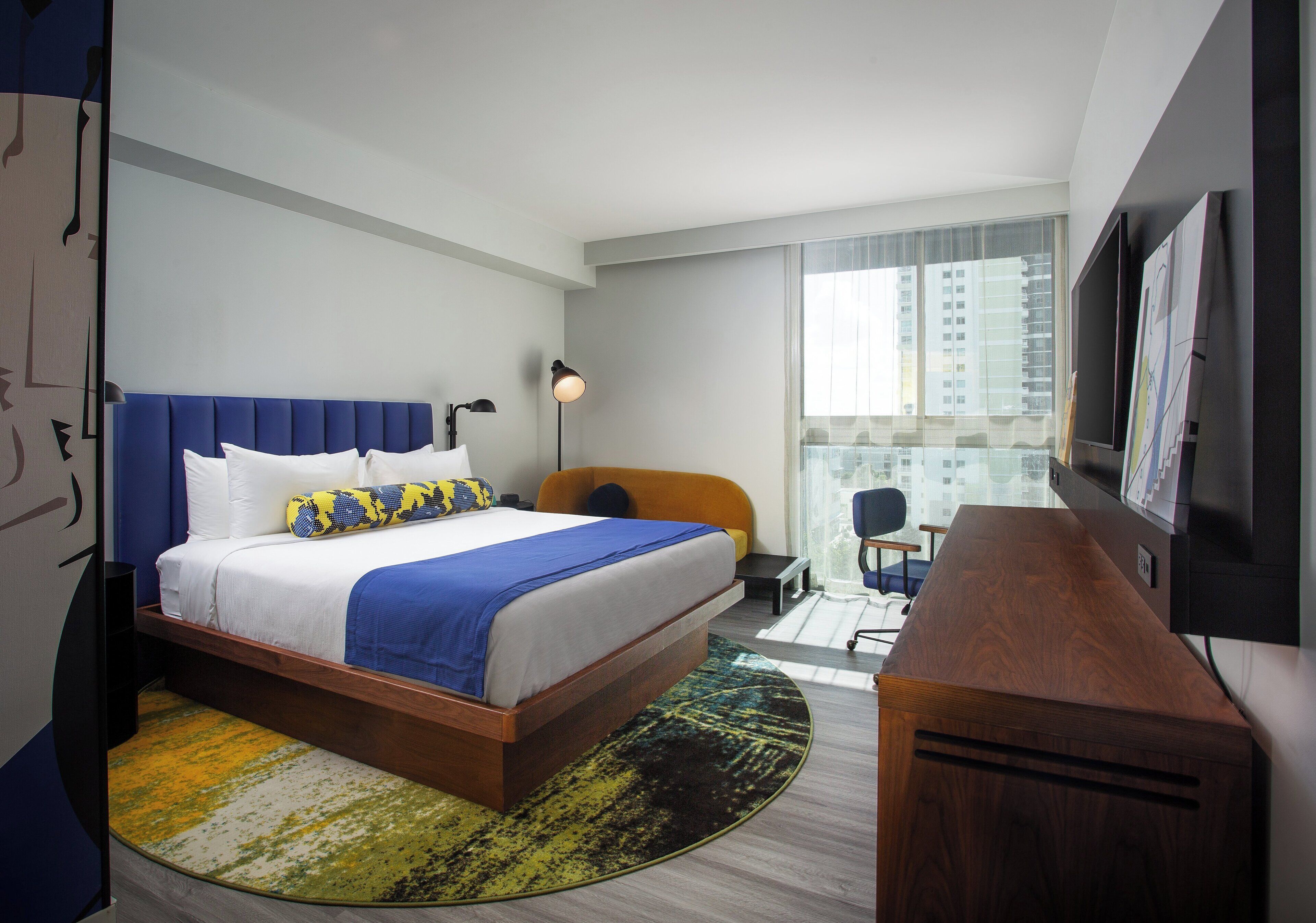 A hotel room with a bed, a desk, a chair, a TV, and a window with a city view.