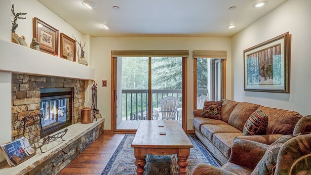 A cozy living room with a stone fireplace, a wooden coffee table, a sofa, and a large window with a view of trees. - Newly Renovated 2 Bed/ 2 Bath Beaver Creek  2 Bedroom Condo by RedAwning (Avon)