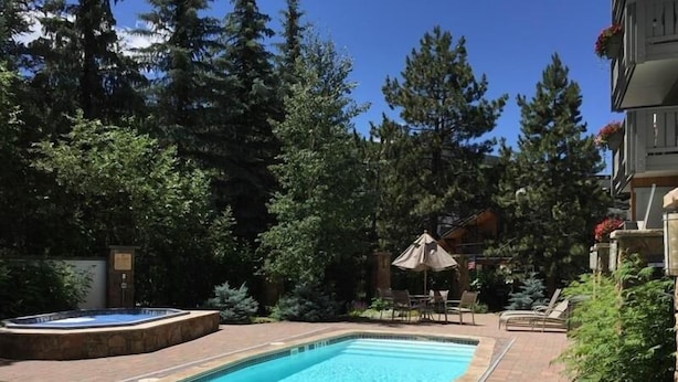 A rectangular swimming pool with a tiled deck, surrounded by trees and a brick pathway. - Nine Vail Road Condos in Heart of Vail Village by RedAwning (Vail)