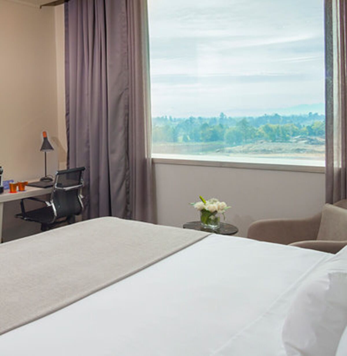 A hotel room with a bed, a desk with a chair, a lamp, and a view of a landscape through the window.