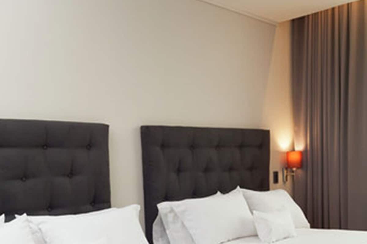 Ein Hotelzimmer mit zwei Betten, jeweils mit dunklem Kopfteil und weißer Bettwäsche. Im rechten Bildbereich ist eine Wandlampe angebracht.