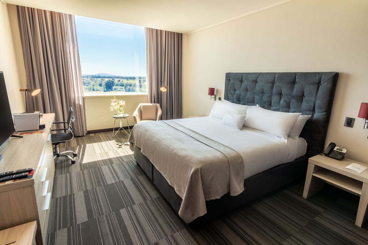 Ein Hotelzimmer mit einem großen Bett, einem Schreibtisch mit Computer, einem Stuhl, einem kleinen Tisch mit einer Vase und Blick ins Freie.