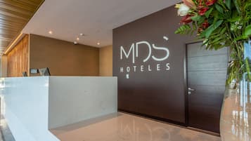 Un área de recepción de diseño moderno, con un mostrador de mármol, una pared oscura con el nombre del hotel y un jarrón grande con flores.