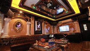 Una sala de karaoke lujosa con un sofá de cuero acolchado, una mesa de vidrio con botellas y una pantalla grande mostrando contenido de karaoke.