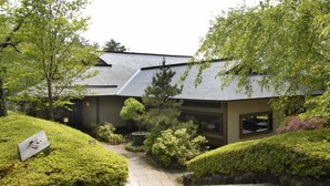 A traditional Japanese-style building with a sloped roof, surrounded by well-maintained greenery and a stone pathway leading to the entrance. - Towa Pure Cottages (Nasu)