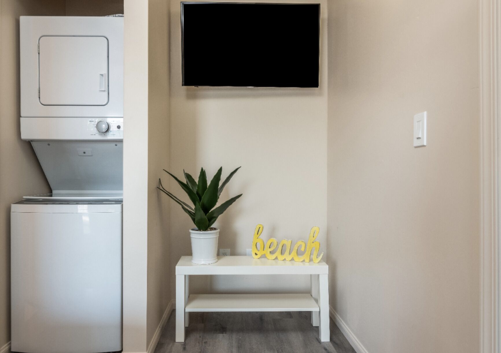A laundry area with a washing machine, a dryer, a small white bench, a potted plant, and a wall-mounted TV.