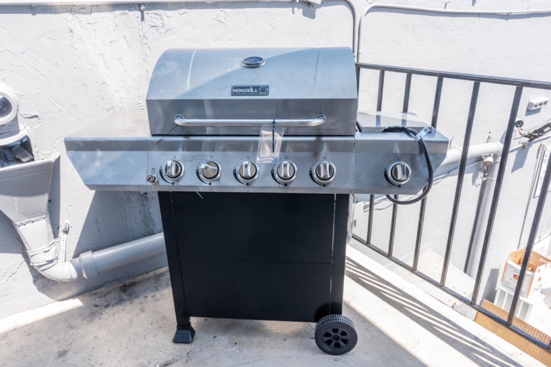 Una parrilla portátil con cinco quemadores, una pantalla digital y un estante lateral, frente a una pared blanca con barandilla metálica.