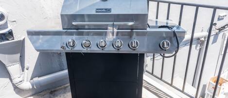 Una parrilla portátil con cinco quemadores, una pantalla digital y un estante lateral, frente a una pared blanca con barandilla metálica.