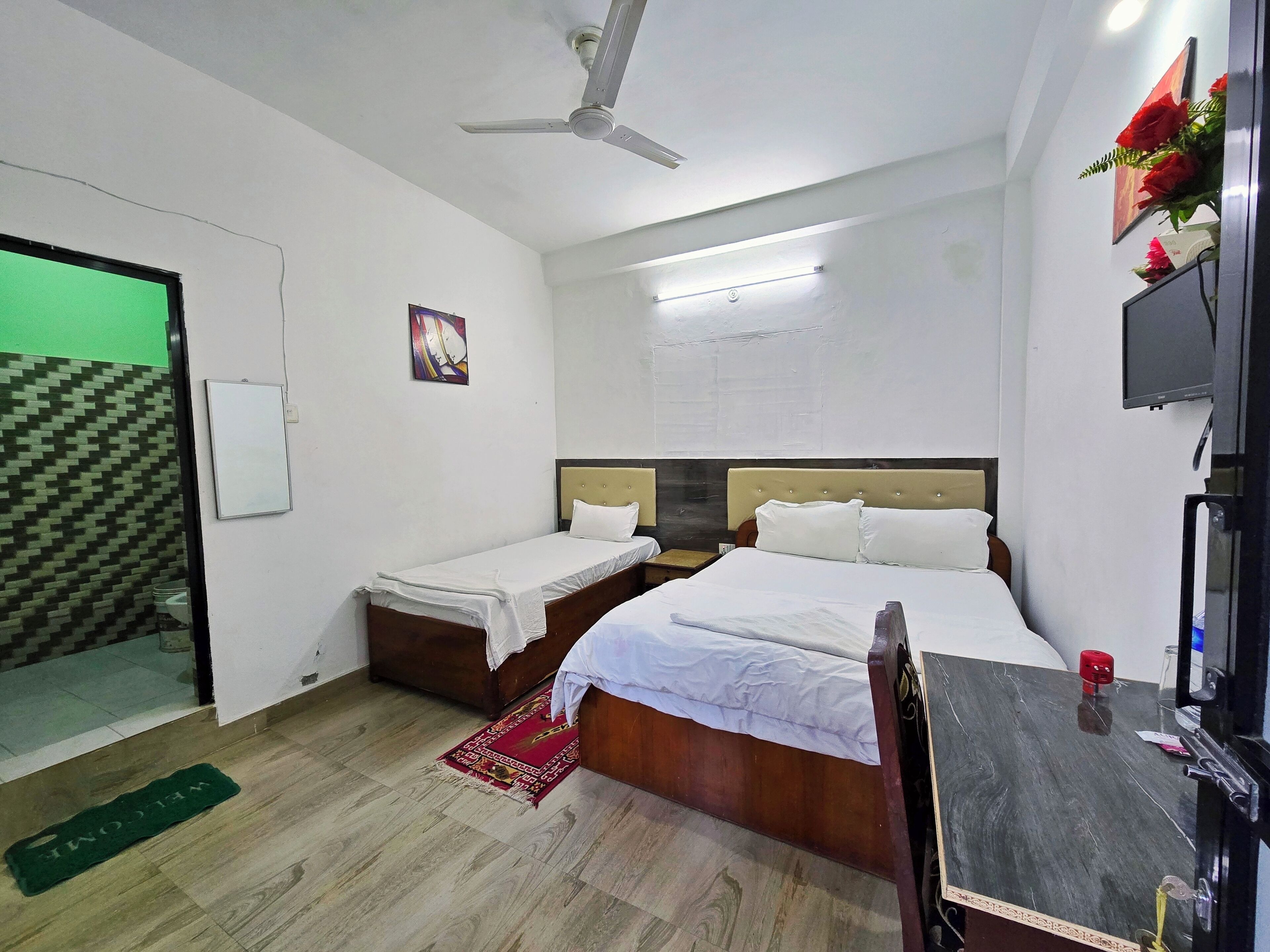 A hotel room with two single beds, a ceiling fan, a flat-screen TV, and a green welcome mat.