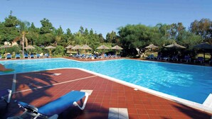 A swimming pool with blue water, surrounded by lounge chairs and umbrellas, and trees in the background. - Studio with air conditioning (Pula Sardinien)